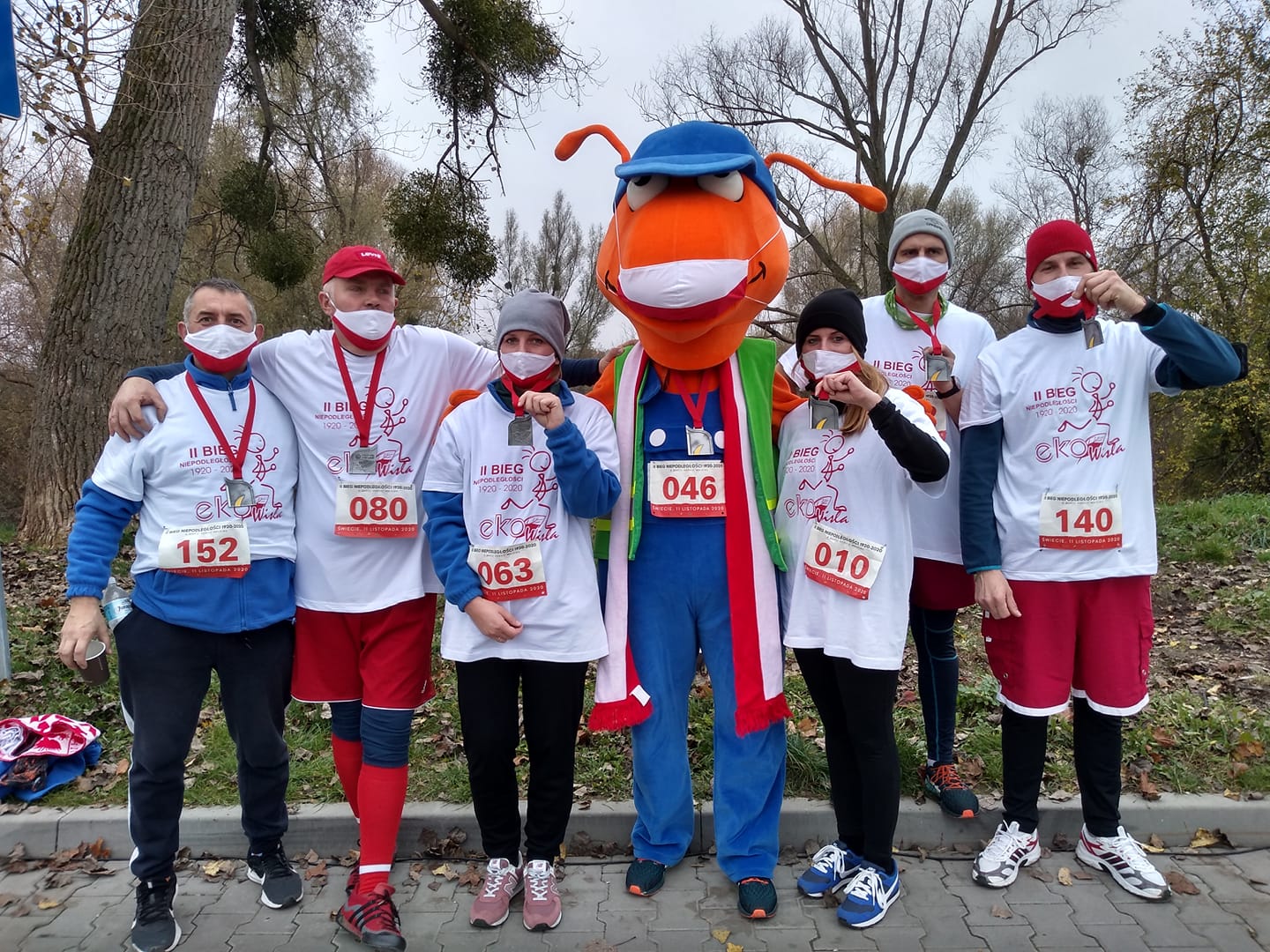 Kostium mrówka i otaczająca go grupa osób stoją na polu, przebrani w kostiumy sportowe i zabezpieczeni maseczkami